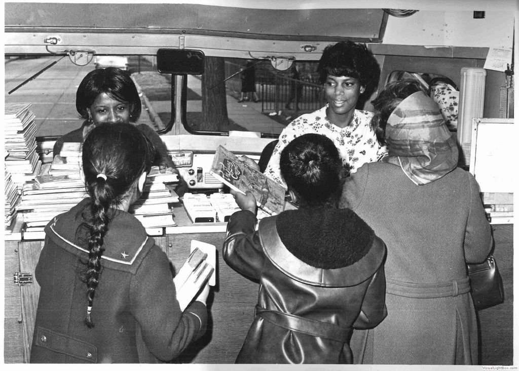 three patrons stand in front of a counter in a bookmobile with two librarians behind the counter