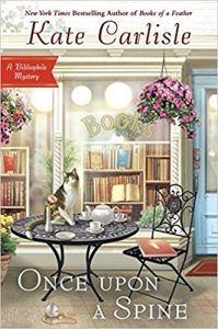 image of sidewalk cafe, table with a cat on it, chair with books, a hanging flowery plant, and a glass storefront window in background with a bookstore behind it