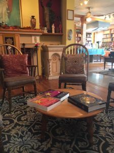 Three chairs surround a low wooden table with large picture books in the foreground, with wooden shelving and books bathed in soft light in the background