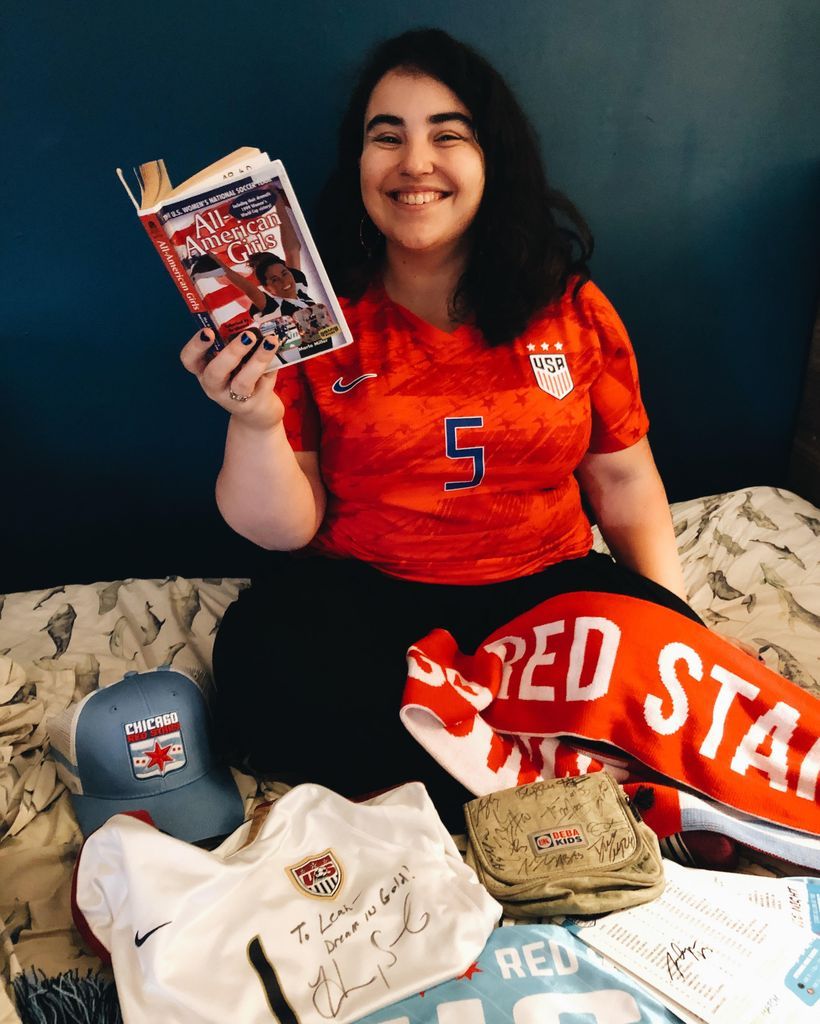 Brunette in a women's soccer jersey reading a book and surrounded by women's soccer gear