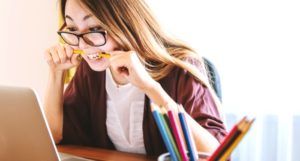 woman biting pencil while sitting on chair in front of computer during daytime