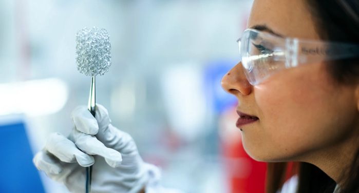 woman scientist in a white lab coat and goggles examining a piece of metallic material https://unsplash.com/photos/HMHt4gAqxP0