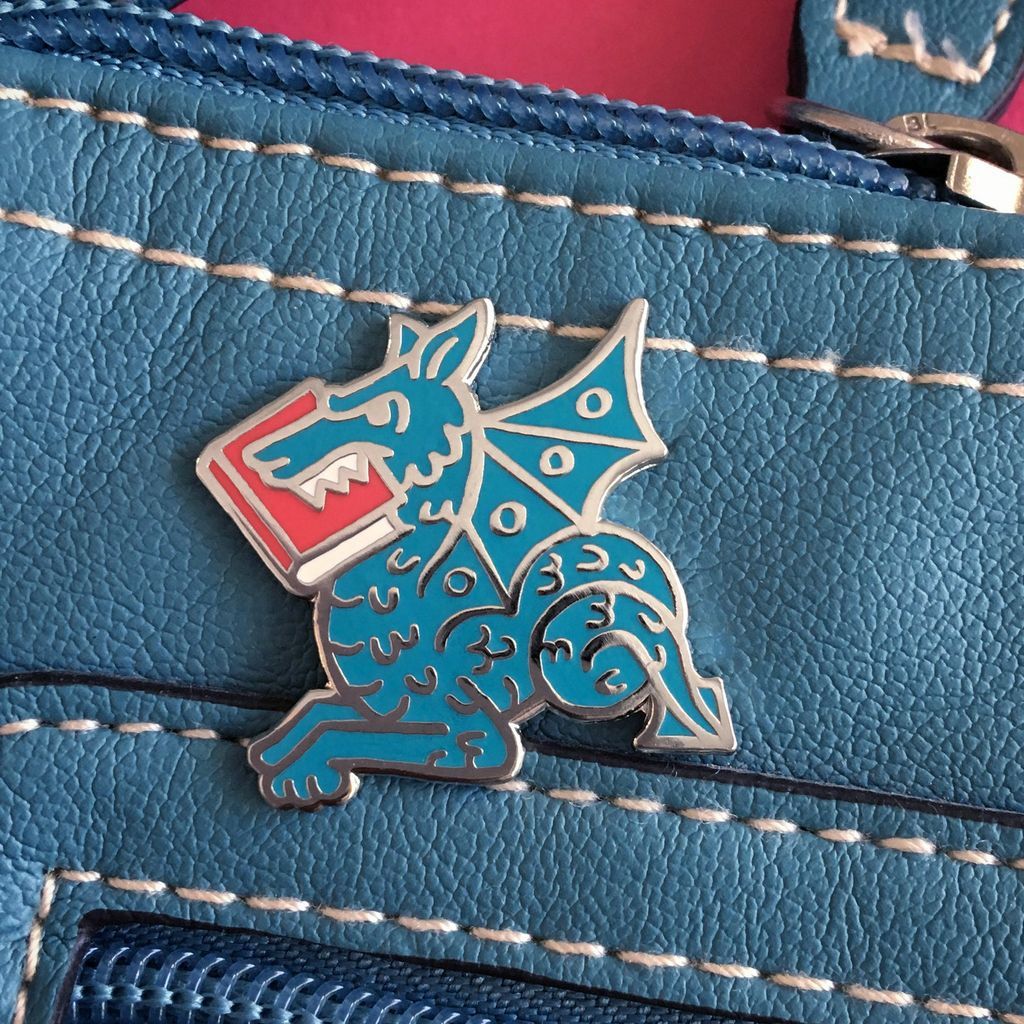 An enamel pin of a medieval-looking dragon with a book in its mouth