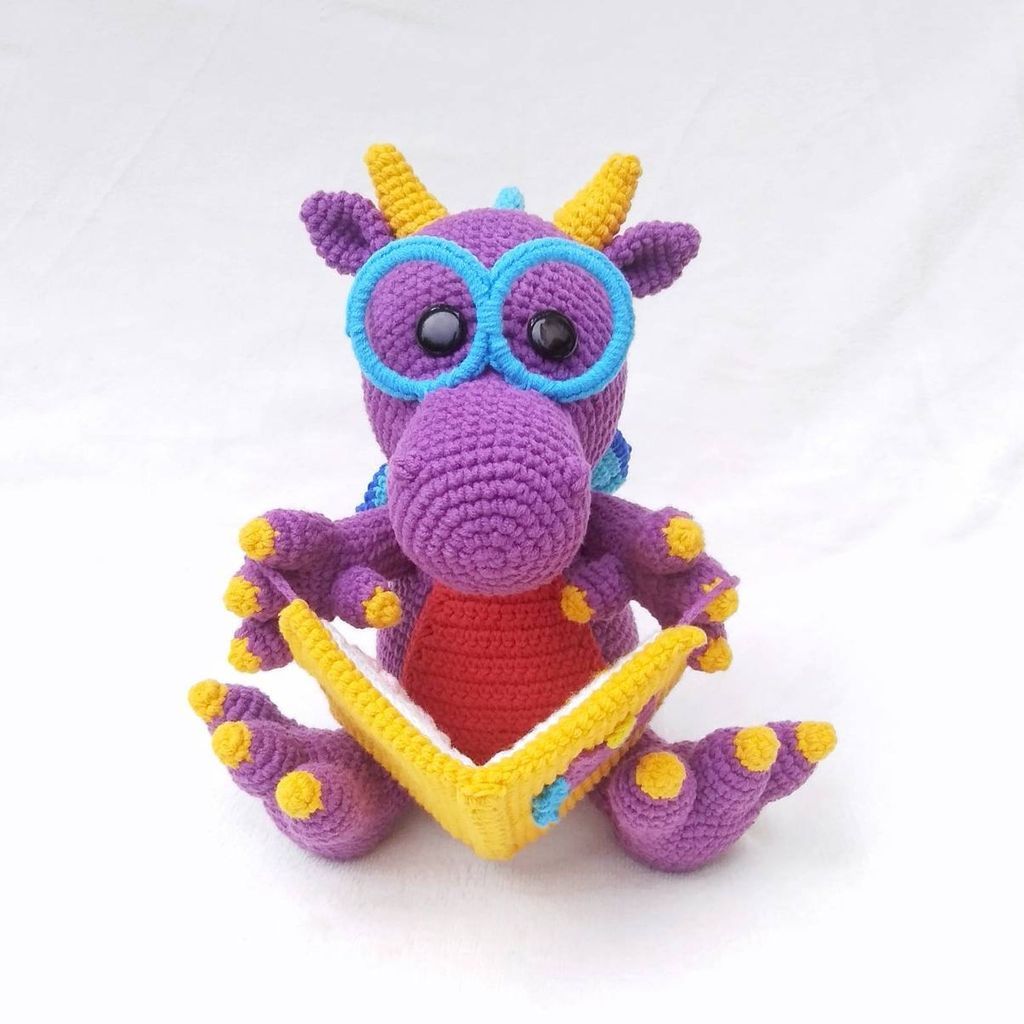 A crochet dragon toy wearing glasses and reading a book