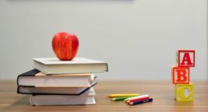 image of an apple on top of a stack of books, some colored pencils, and letter blocks on a wooden surface https://unsplash.com/photos/OyCl7Y4y0Bk