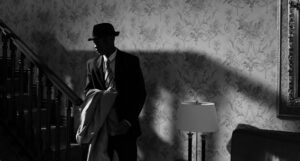 black and white photo of a man in a fedora holding a trench coat at the bottom of a staircase in a wallpapered room