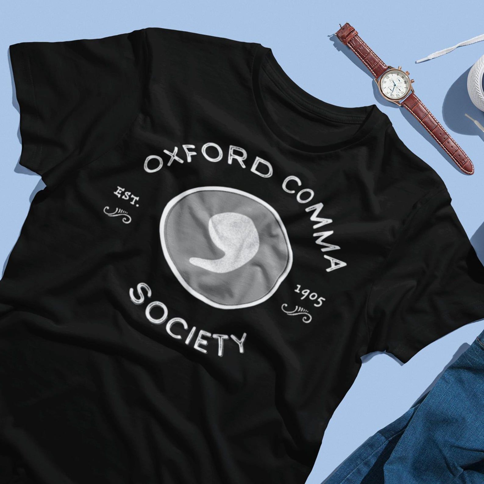 Black, Oxford Comma Society t-shirt laid out on a blue background