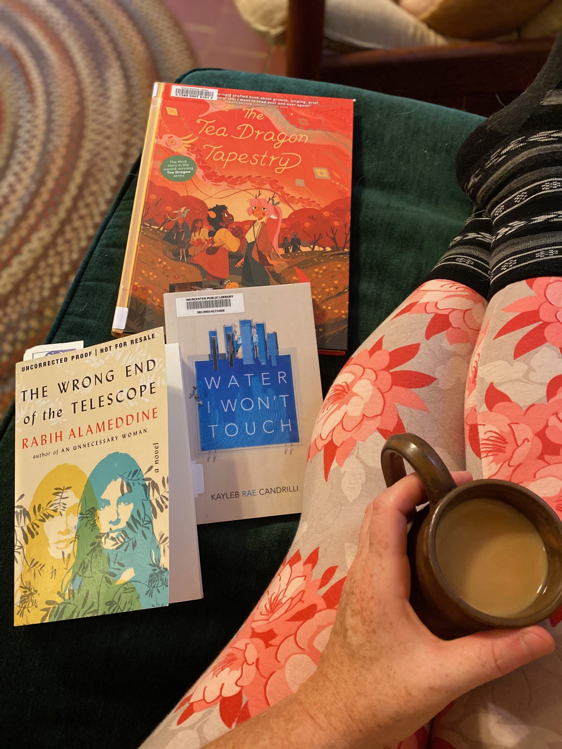Image of my legs stretched out on a green couch. I am wearing leggings with large red flowers on them and holding a mug of tea. Three books ar on the couch next to me: The Tea Dragon Tapestry, Water I Won't Touch, and The Wrong End of the Telescope. Photo by me.