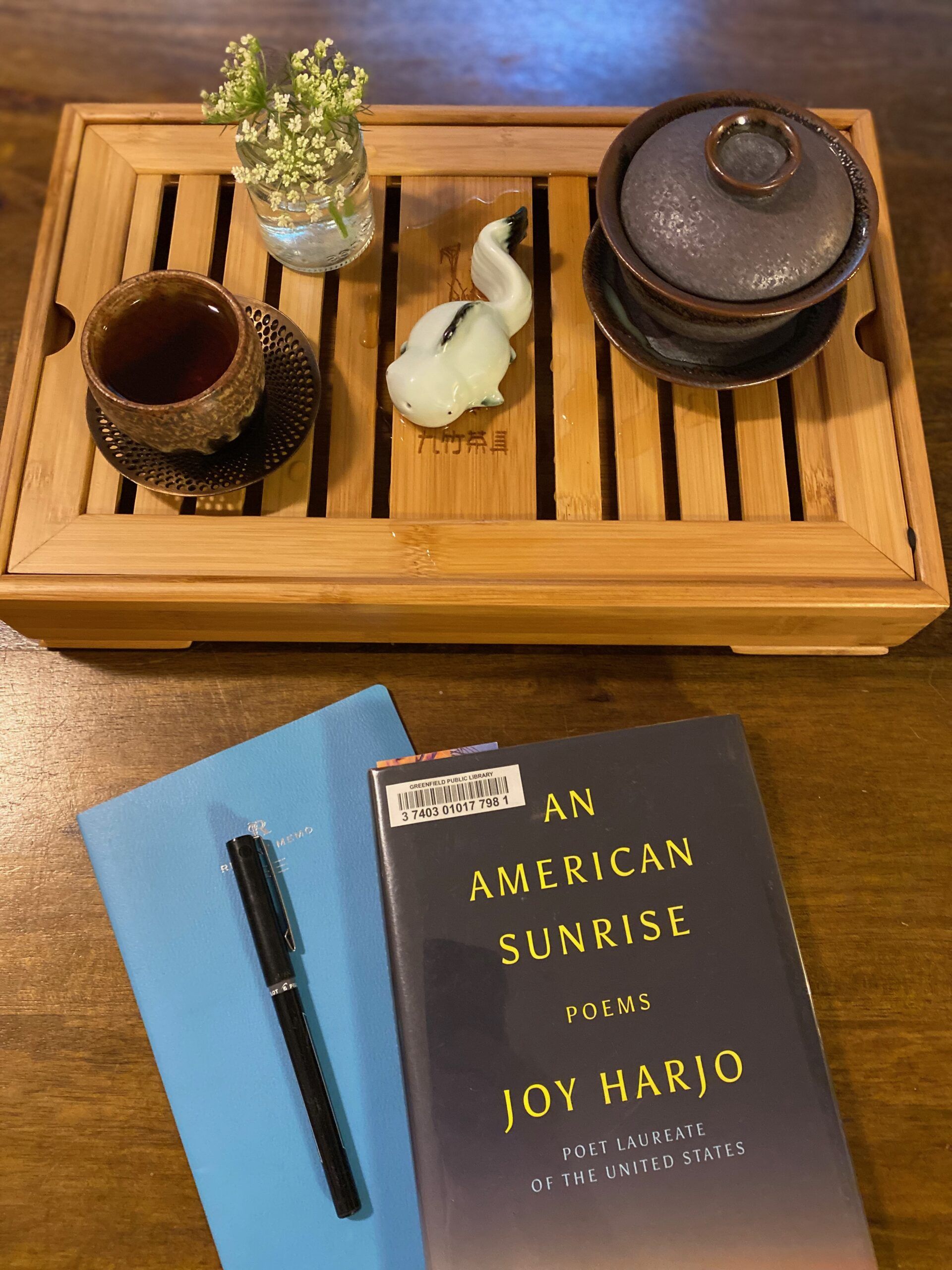 A tea setup on a wooden tray with open slats: a small ceramic mug on a metal coaster, a goldfish tea friend, a small jar of Queen Ann's lace, and a small Gaiwan teapot. A blue notebook and An American Sunrise by Joy Harjo sit on the table in front of the teaware. Photo by me.