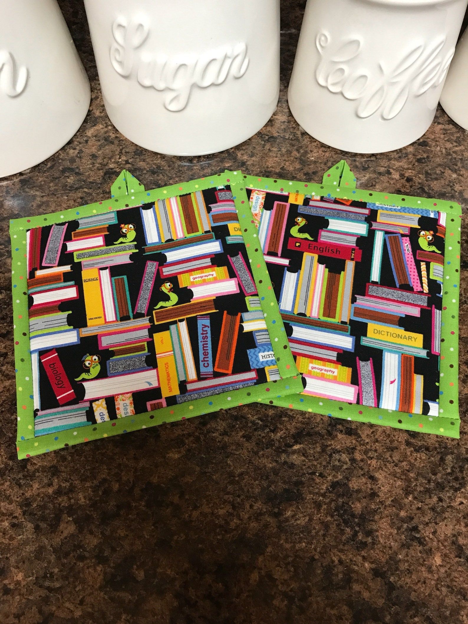 On a brown marble kitchen counter, there are two potholders. They are both black with an array of different books and a green multi-colored polkadot edges. There are also three bookworms between the piles of books. 