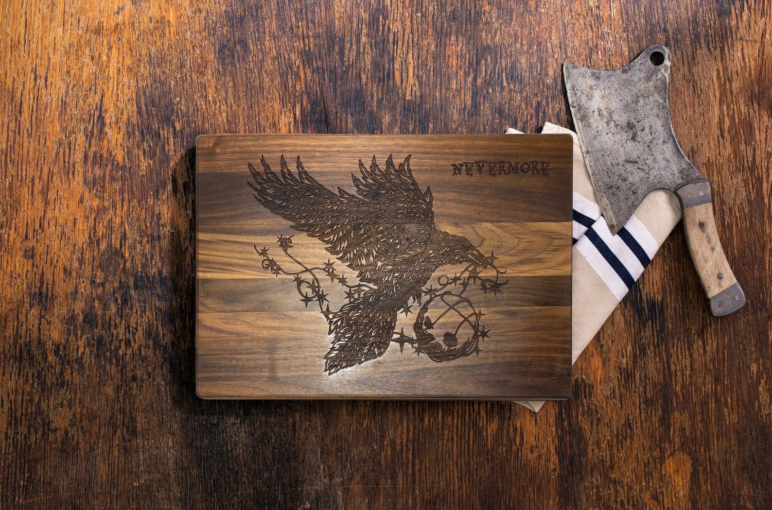 An overhead shot of a wooden charcuterie board. Etched into the wood is a graphic of a raven with the words, "Nevermore" in the top right hand corner. To the right of the cutting board are two striped tea towels and a butcher knife.