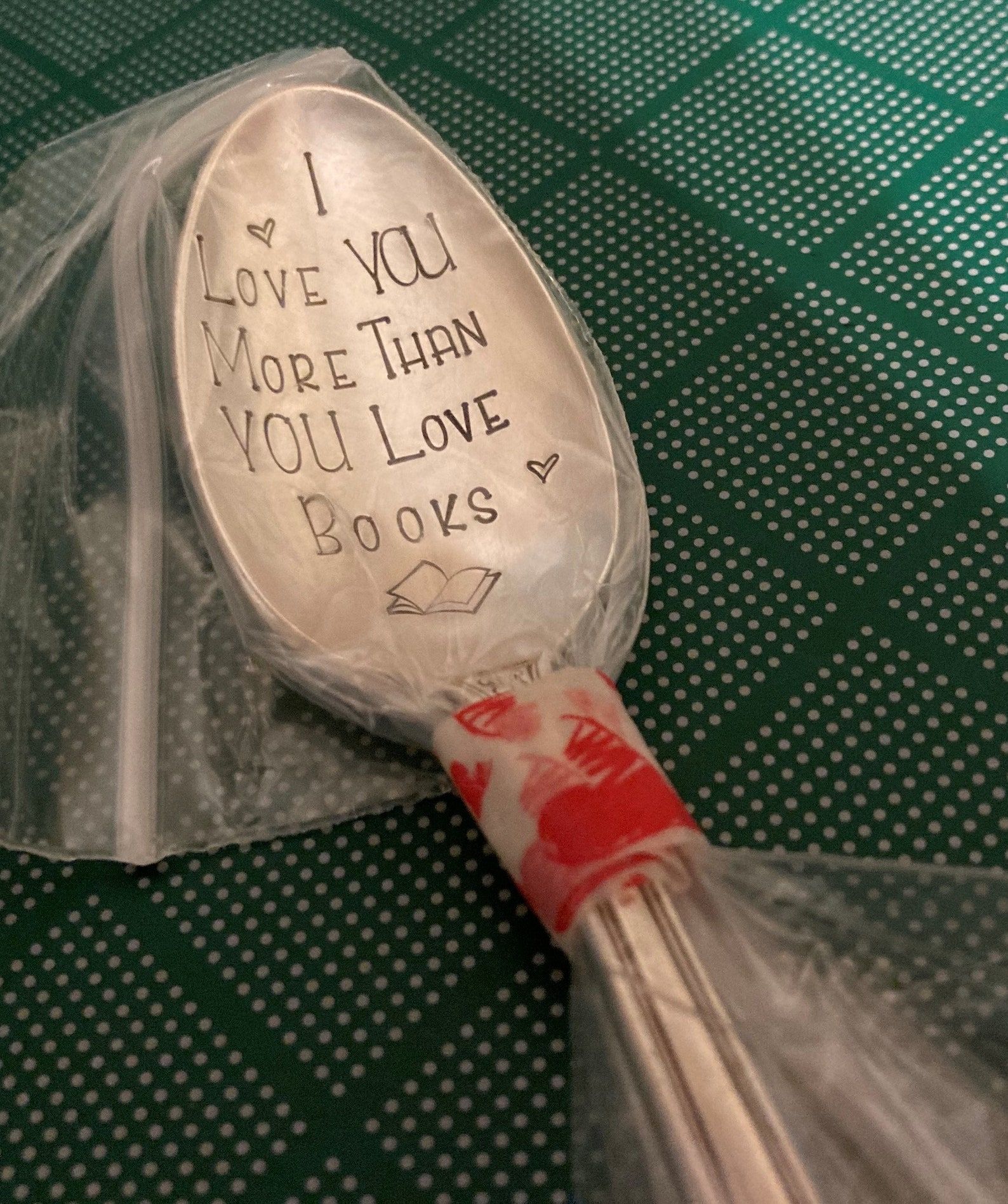 An engraved silver spoon lays on a green polkadot mat. The spoon is wrapped in plastic but the engraving is still visible, it reads, "I love you more than you love books."