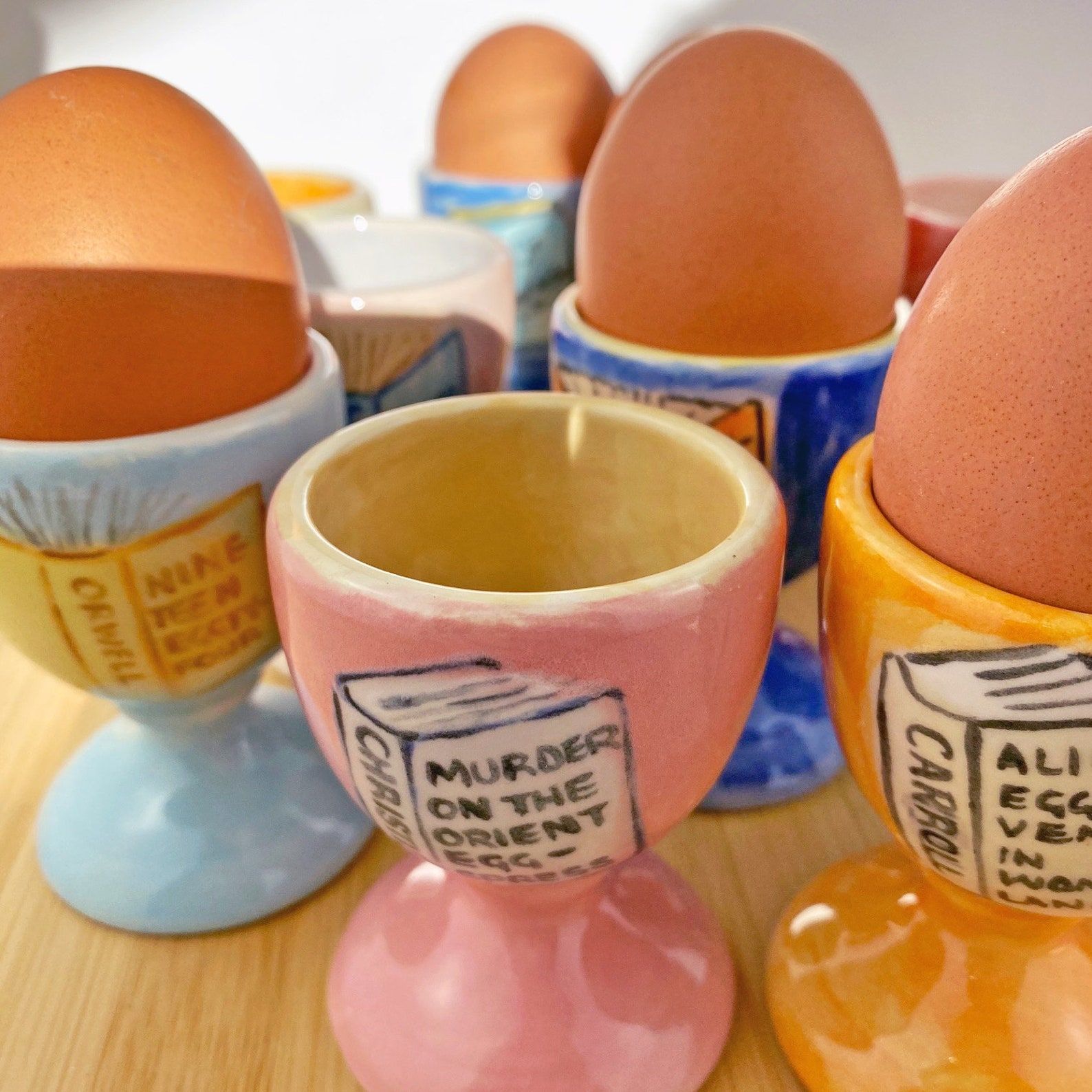 A photo of a group of painted eggs with egg-related book title puns. The egg cups are different colors and hand-painted.