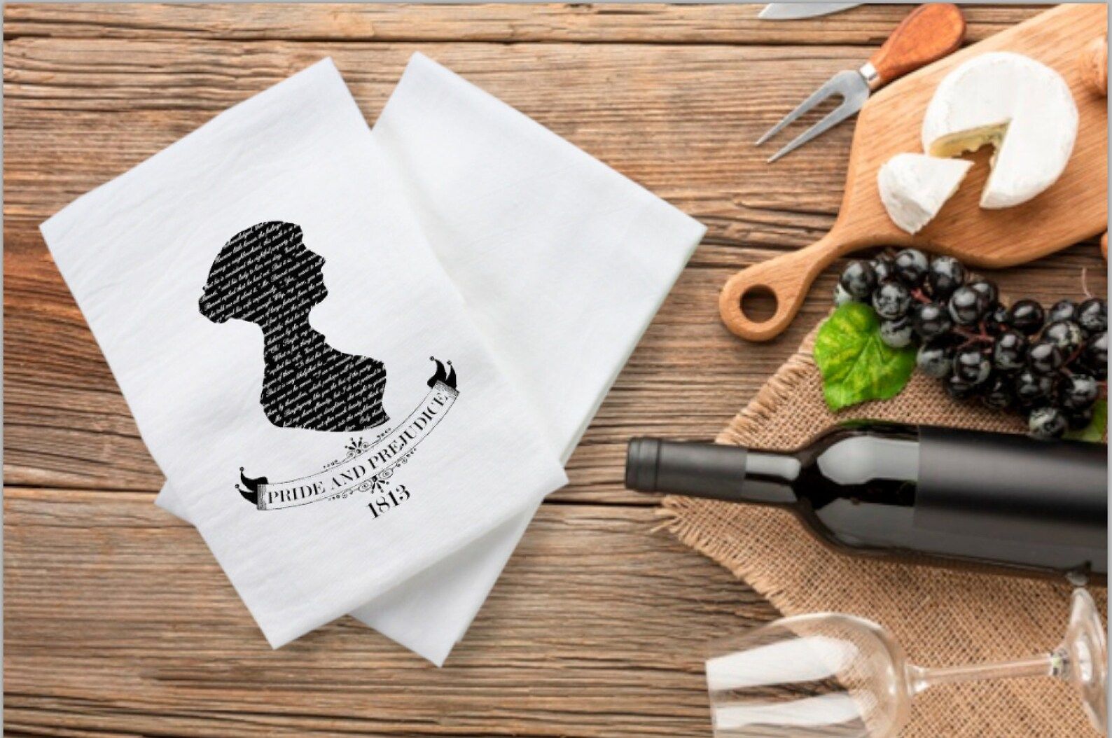 An overhead photo of two white tea towels with a silhouette of a woman in black and the title, "Pride and Prejudice" with the date 1813 below it. To the right of the tea towels is a bottle of wine, glasses, grapes, and a cheese board.