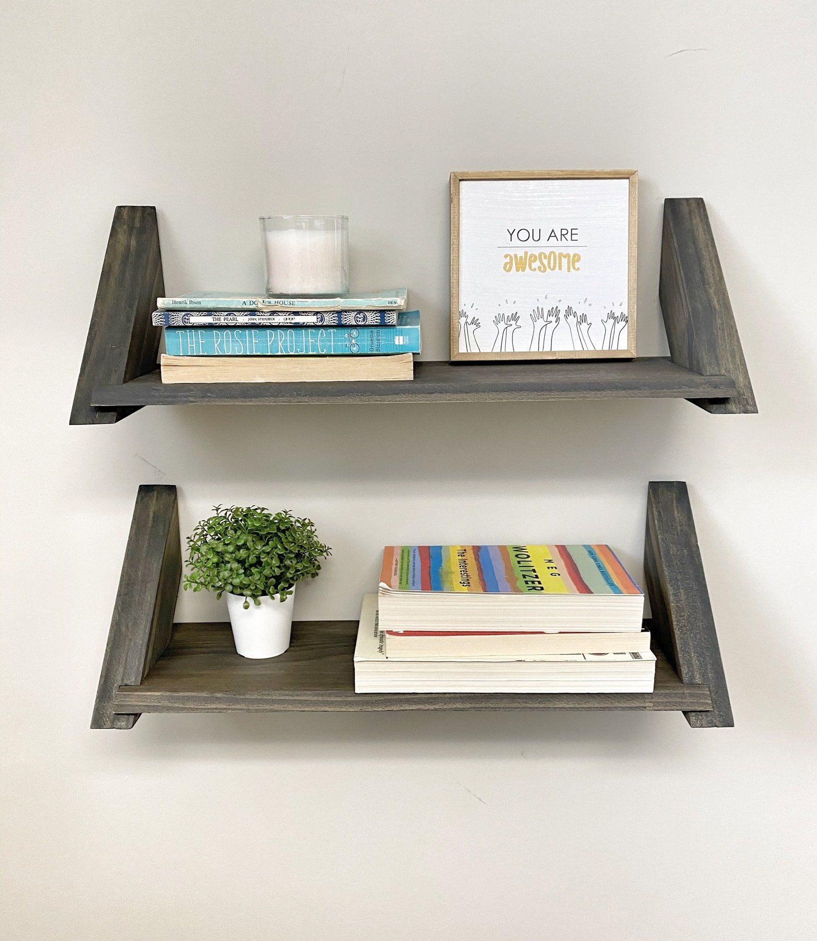 two hanging gray wooden shelves with books, plants, and candles on the shelves