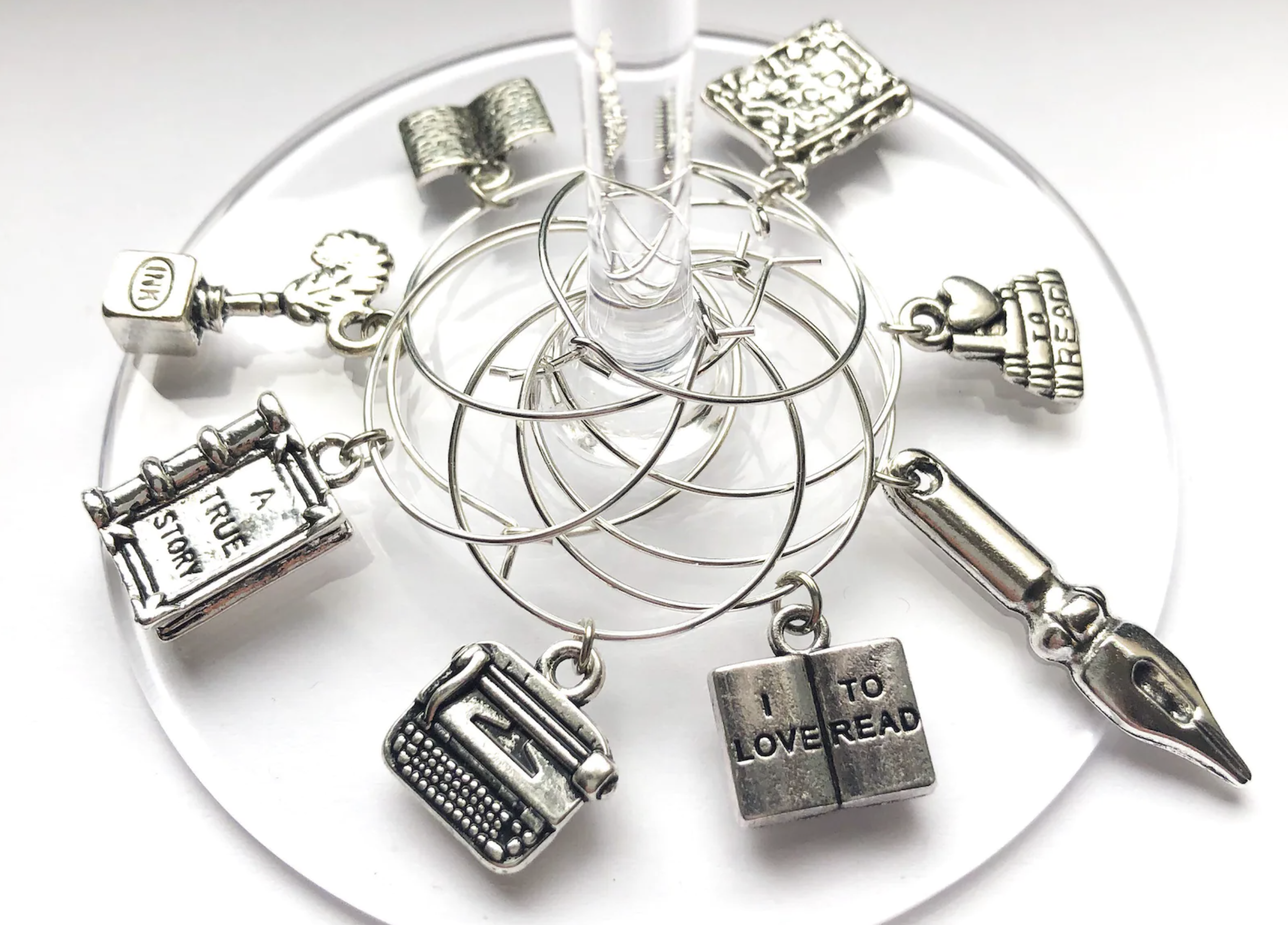 Eight wine glass charms are looped around the stem of a clear wine glass. The charms are silver and in the shapes of books, pen nibs, a typewriter, and a beanie hat. 