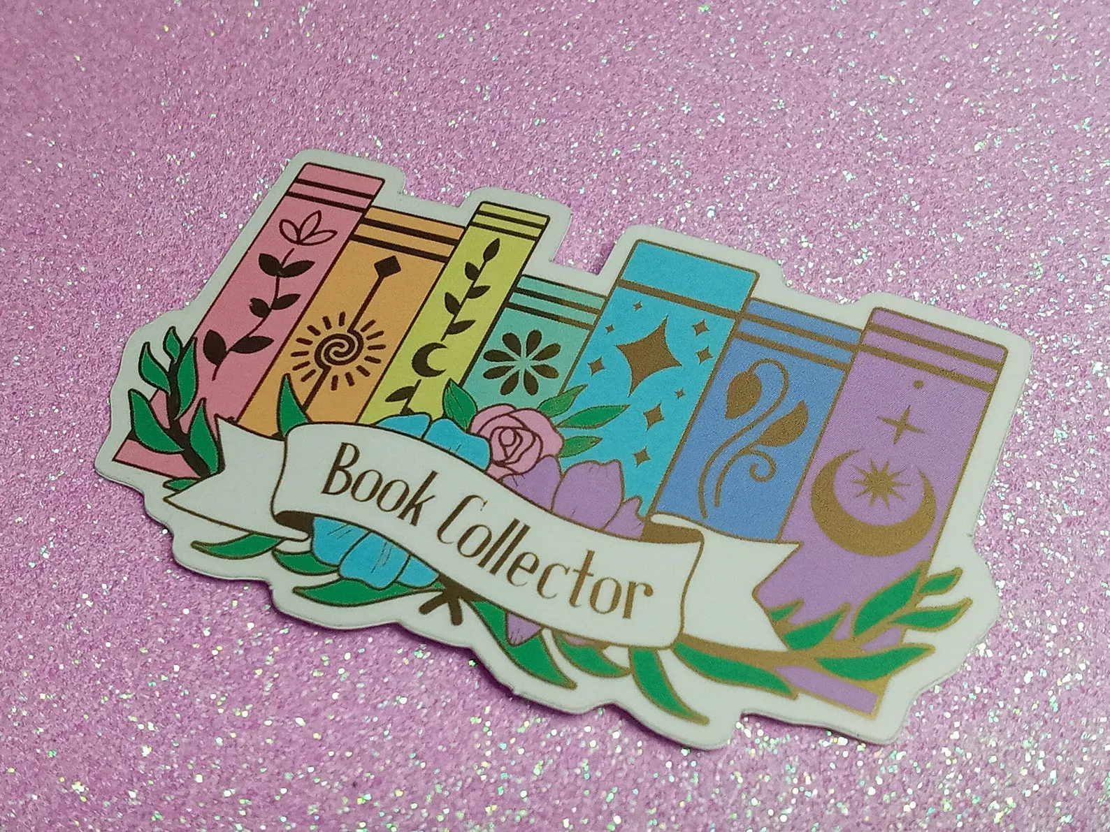Image of a sticker on a glittery purple background. The sticker has six rainbow-colored book spines, each with a fun gold design. There is a banner reading "book collector" in front of the books. 