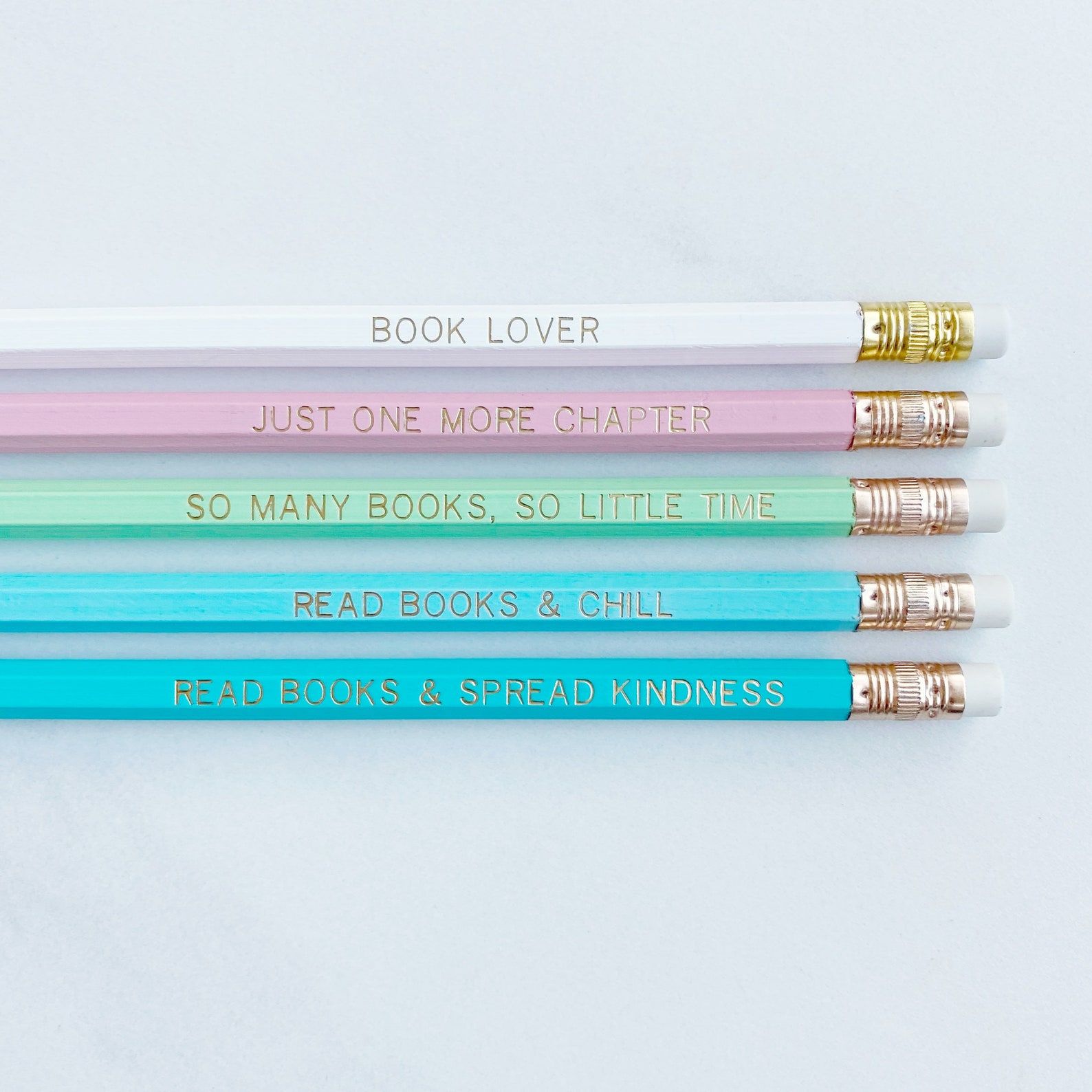Image of five pencils in various blue shades, as well as green, pink, and white. The pencils each have a bookish phrase in gold text. 