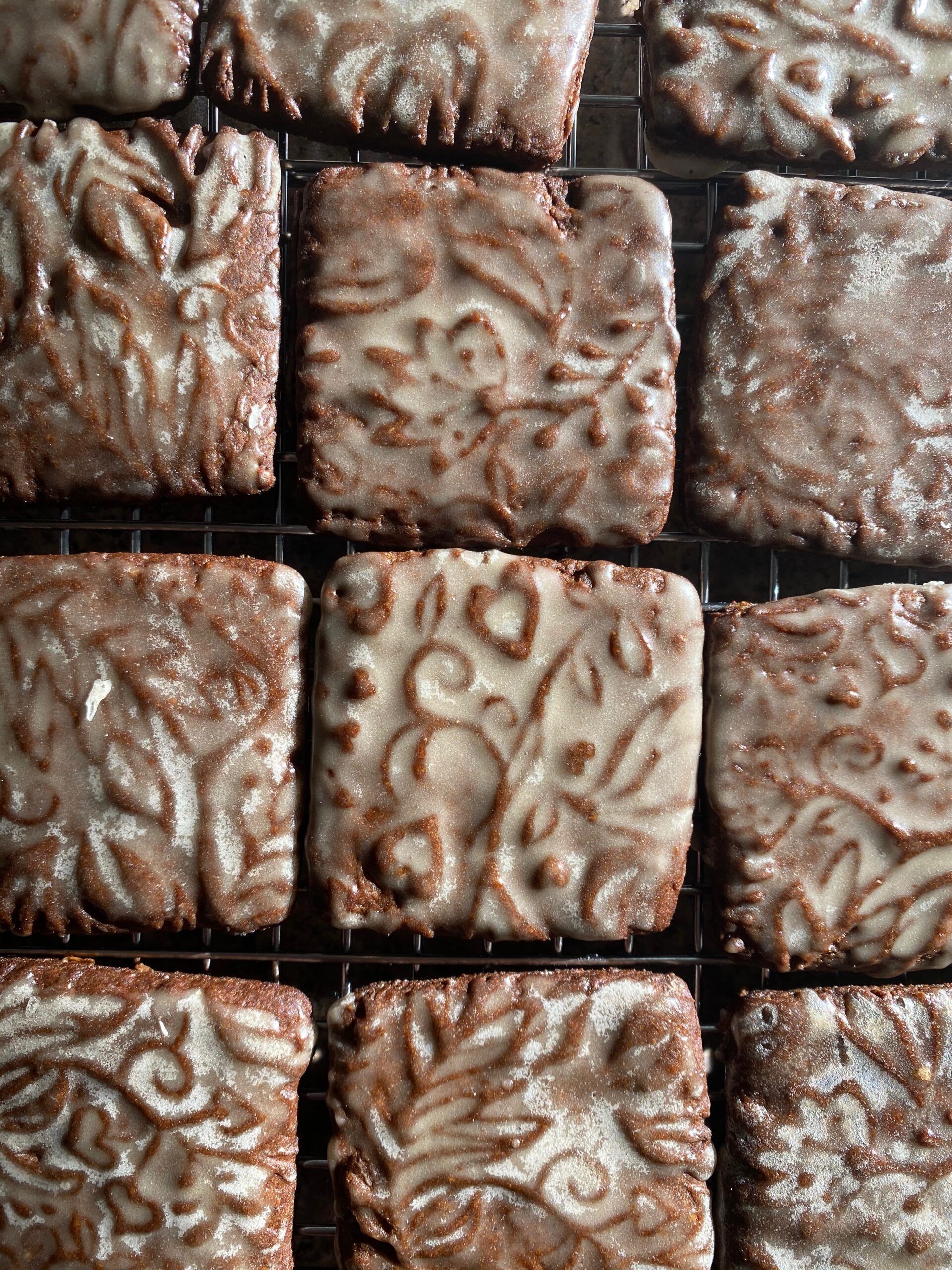 Several small square gingerbread cookies, imprinted with a pattern of leaves and flowers and covered in a shiny white glaze.