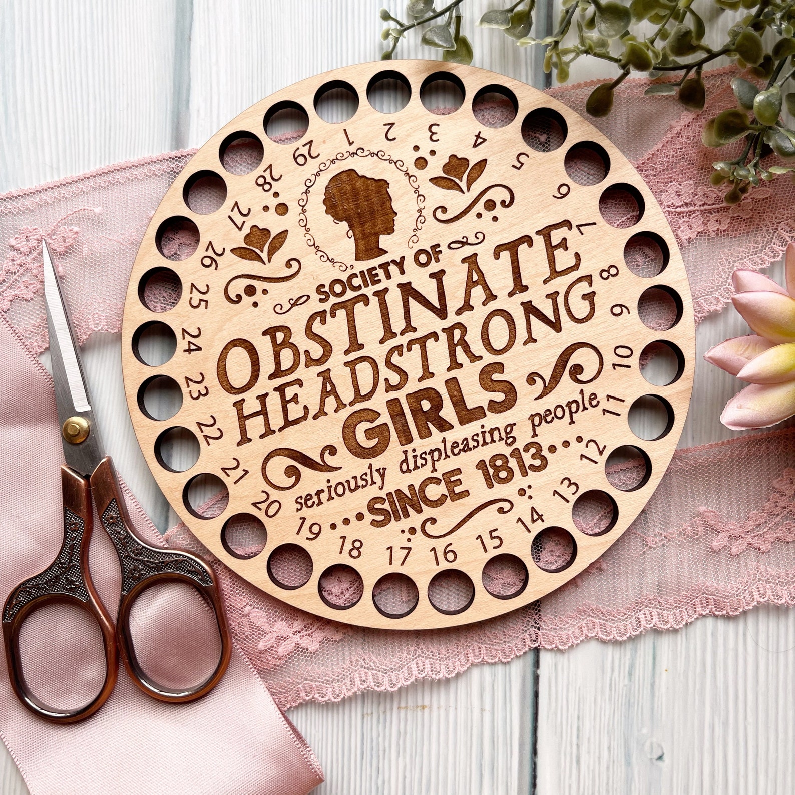 A wood circular thread organizer with "Society of Obstinate headstrong girls" embossed on it.