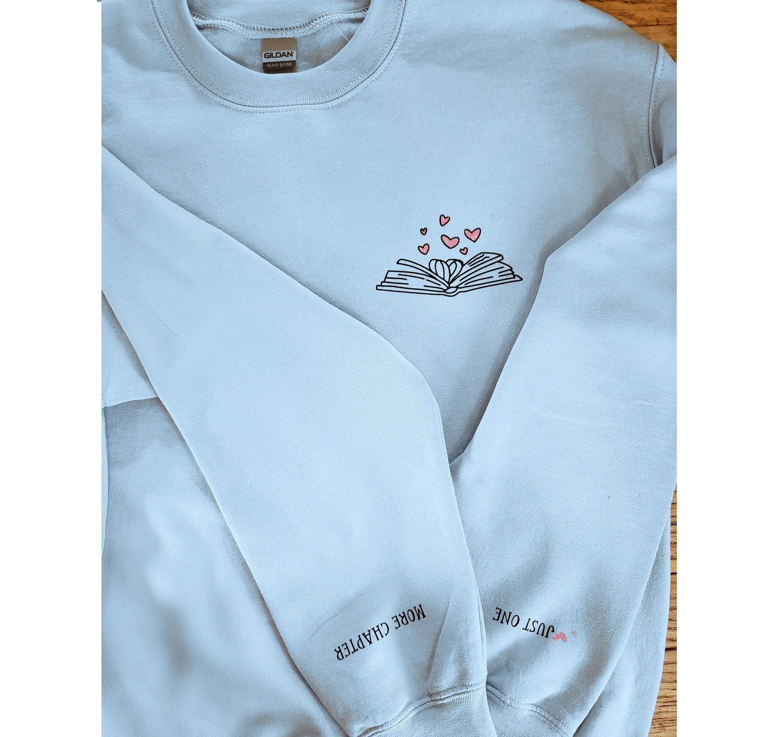 Image of a white sweatshirt. It has an open book in black ink that's open, with pink hearts above it. On the sleeves are the words "just one" and "more chapter." 