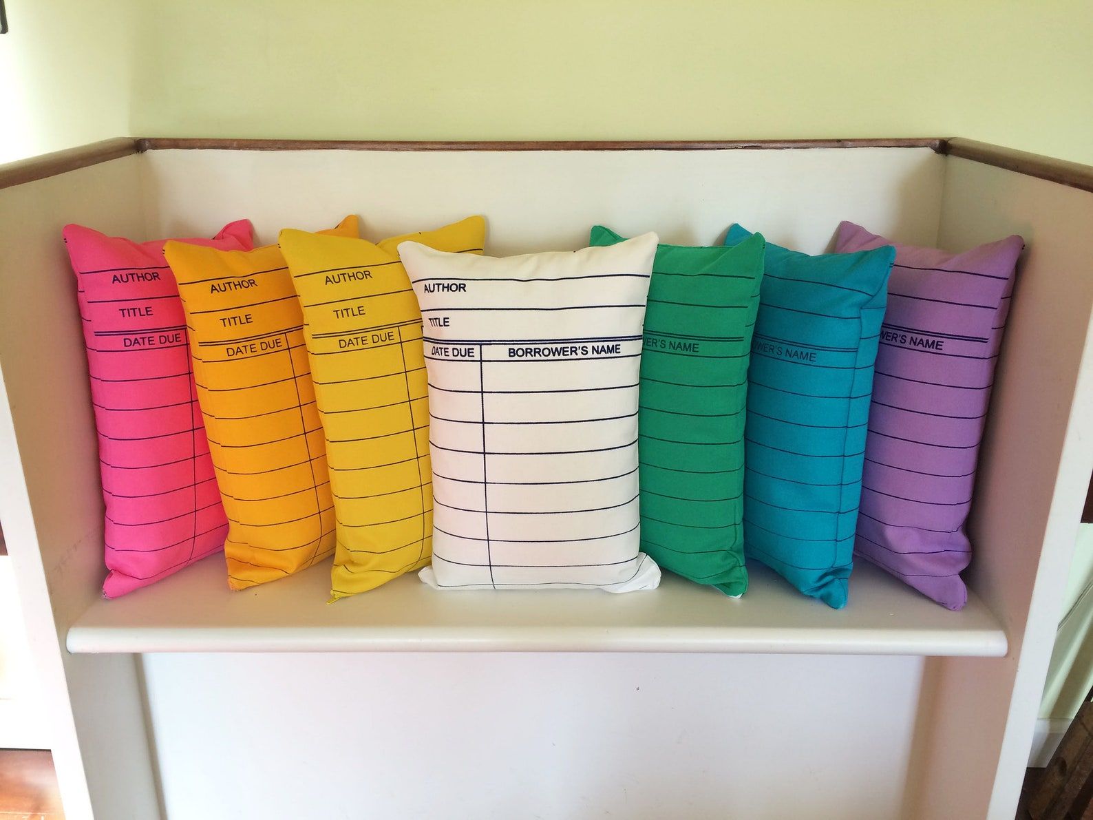 Image of seven colorful pillows, all in the shape of a library due date card. 