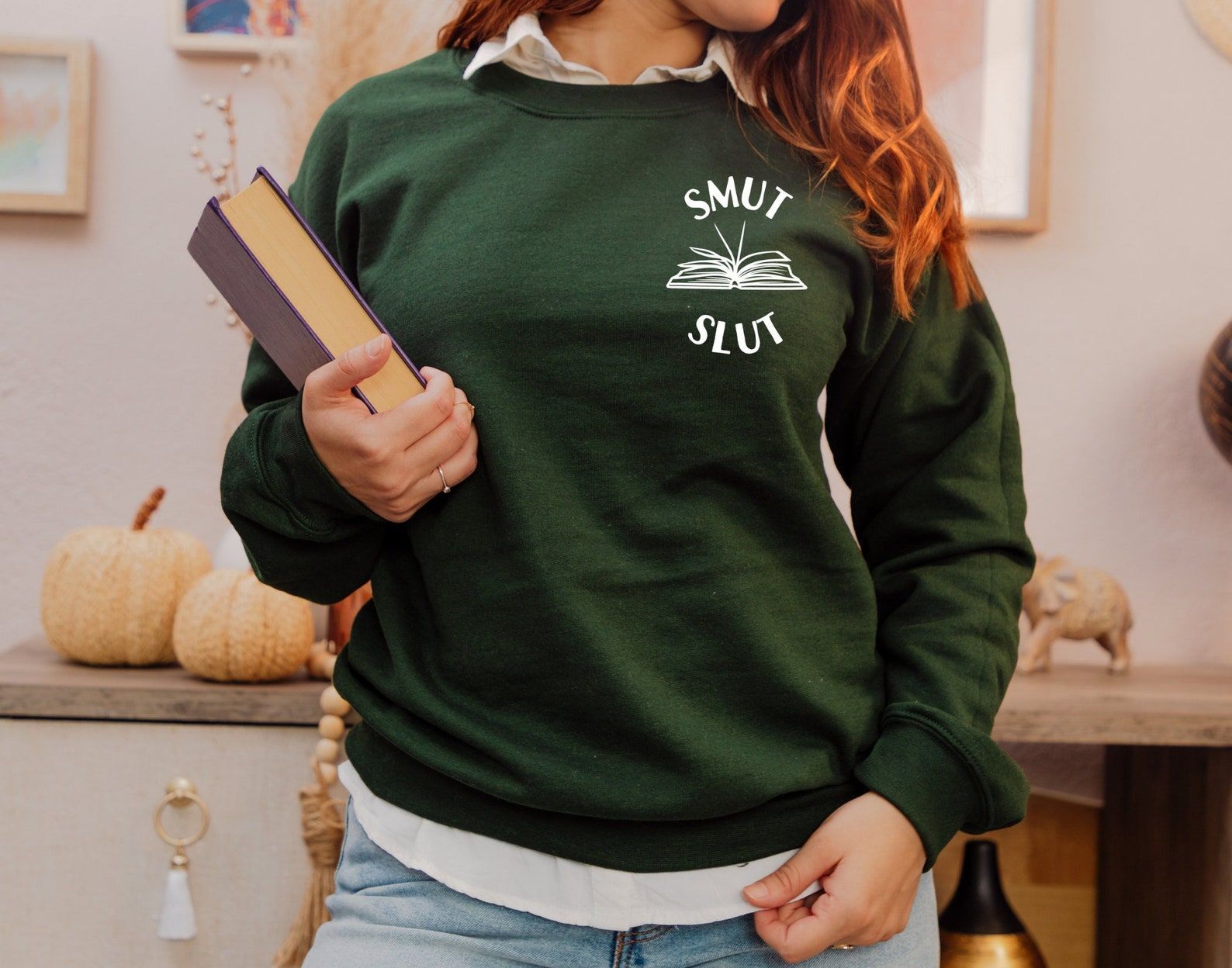 Image of a person with white skin and long red hair wearing a green sweatshirt. In white ink at the chest are the words "smut slut" and an open book. 