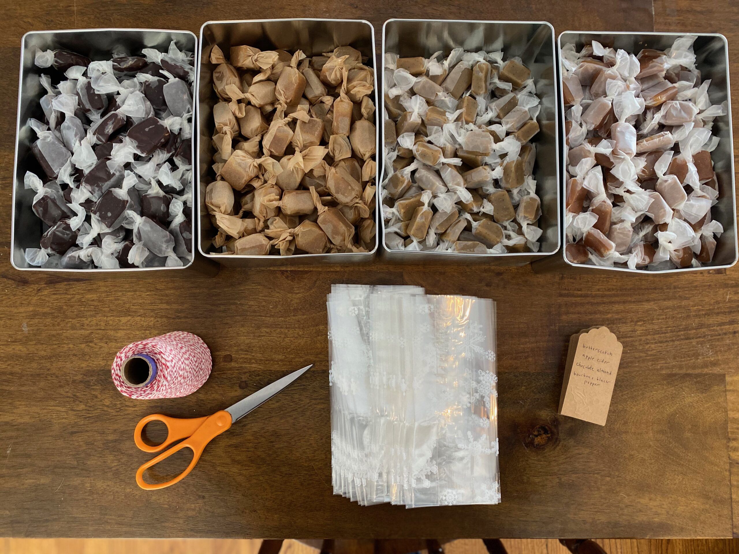 Four square tins of homemade caramels, different colors, wrapped in wax paper. A bolt of red and white twine, a pair of scissors, brown gift tags, and a stack of small decorative bags sit on the table in front of the caramels. Photo taken by me.