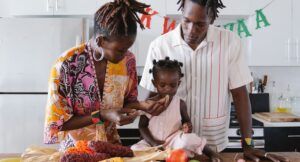 a photo of a family celebrating Kwanzaa. Two Black parents are feeding their toddler fruit with a Kwanzaa banner in the background.