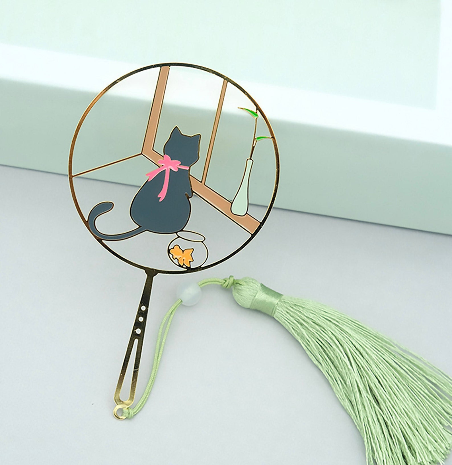 Image of a bookmark featuring a black cat and goldfish. It's a thin, round bookmark with a green tassel. 