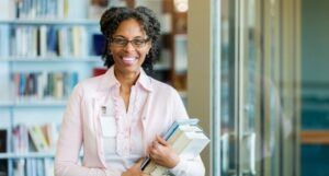 portrait of a mature Black woman standing in a library holding a stack of books