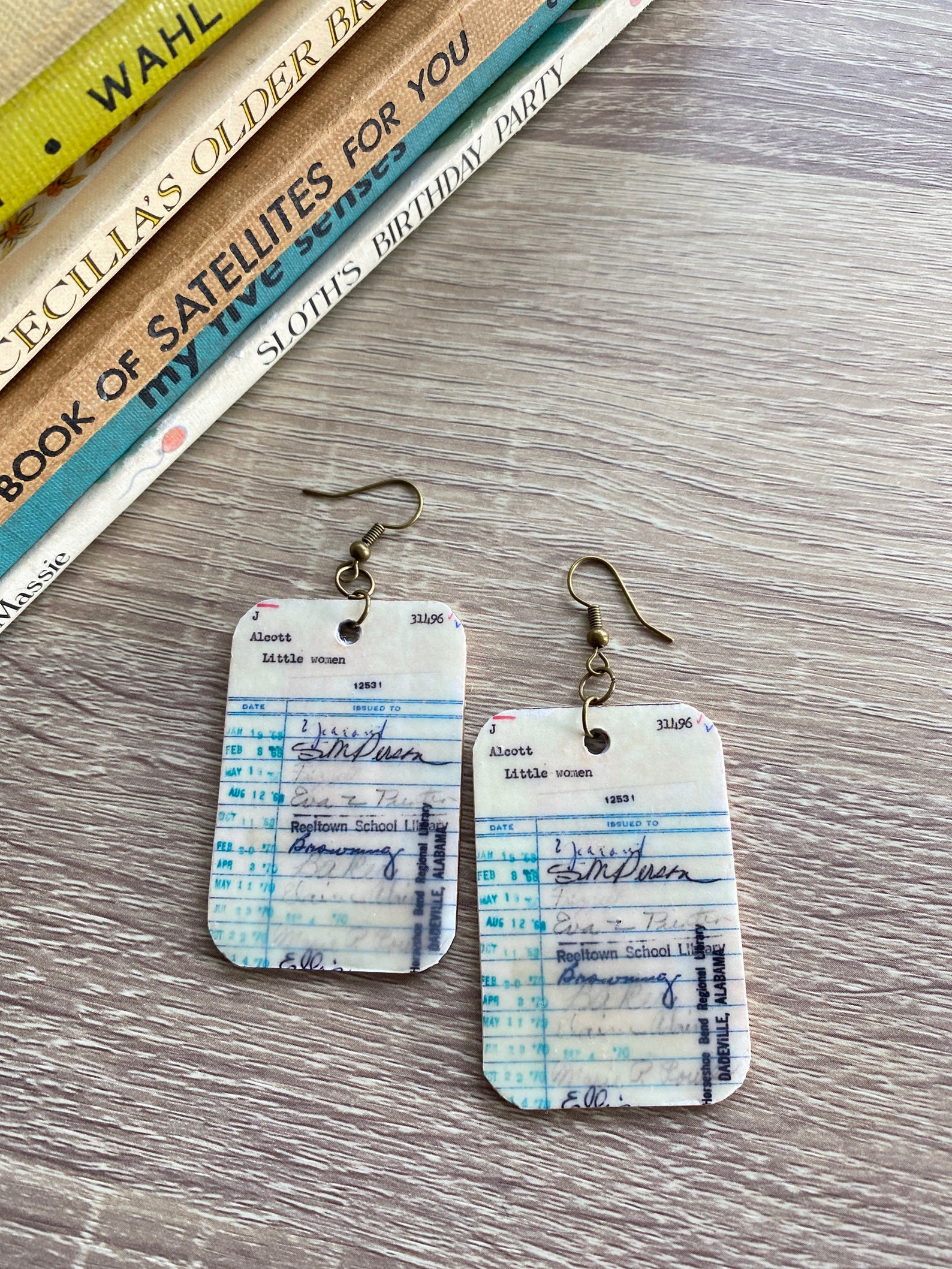 Image of two earrings designed like a library checkout card for the book Little Women. They're on a table. 