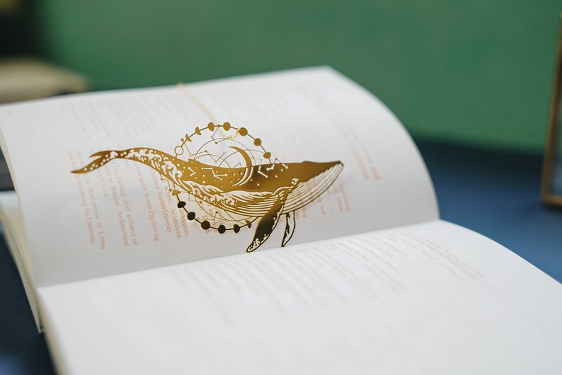 Image of a whale bookmark. The whale has celestial scenes and the moon phases behind it. 
