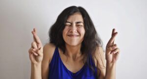 woman in a blue tank top with her eyes closed and fingers of both hands crossed