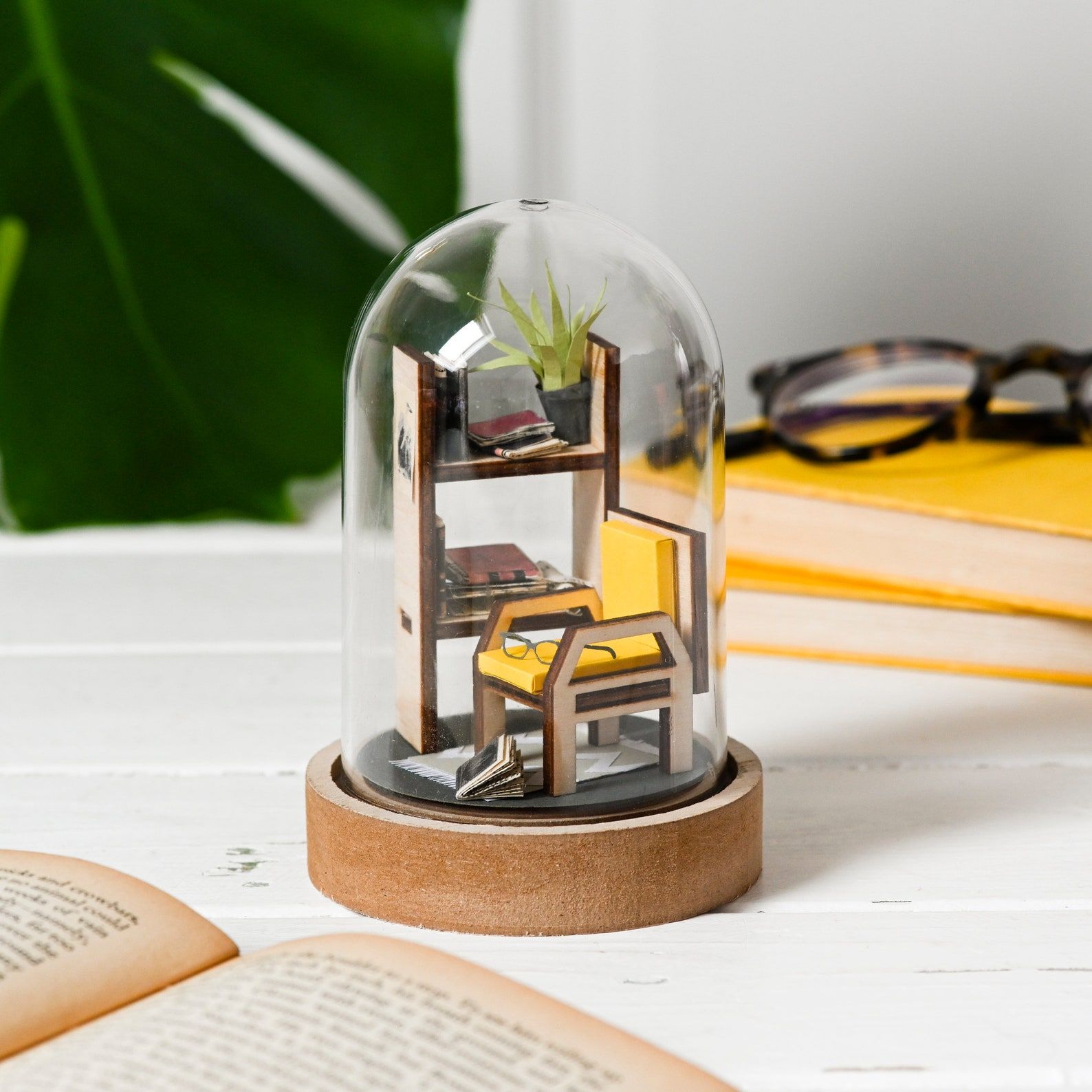 Image of a paper sculpture inside a display jar. The paper sculpture has a yellow chair, glasses, and books on a shelf. 