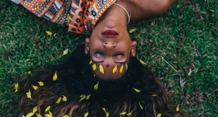 a brown-skinned Indian person with long hair, a red bindi, and red lipstick lays down on grass with their eyes closed. There are yellow petals in their hair