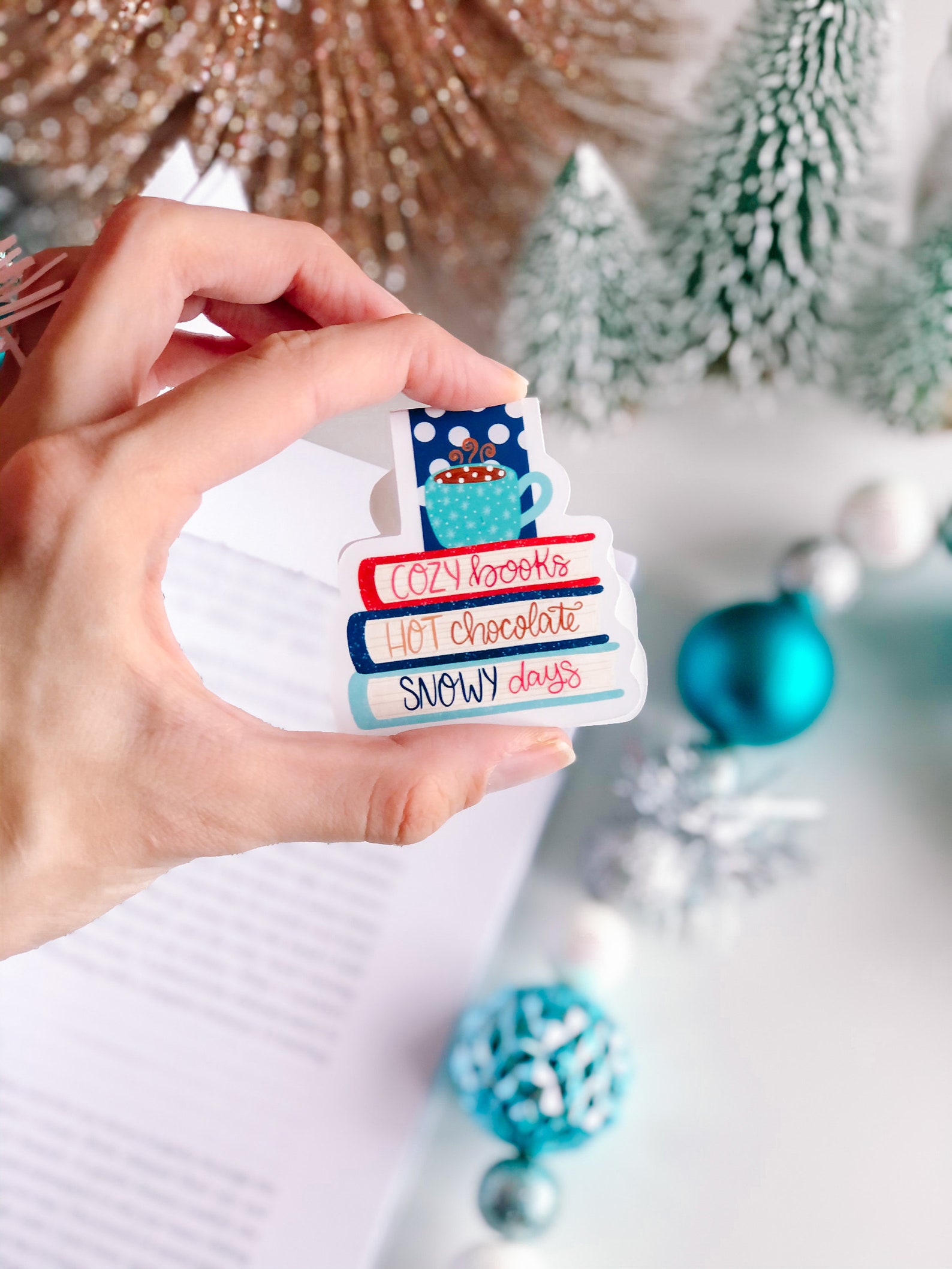 A magnetic bookmark depicting a colorful stack of books and a steaming cup of hot cocoa that reads "Cozy books, hot chocolate, snowy days"