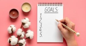 Image of a notebook with the word "goals" on a pink background. A white hand holds a pen.