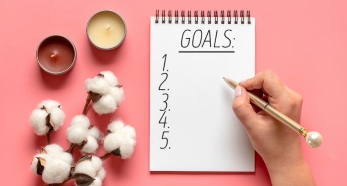 Image of a notebook with the word "goals" on a pink background. A white hand holds a pen.