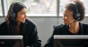 two brown skinned people working at computers with headsets on looking at one another affectionately