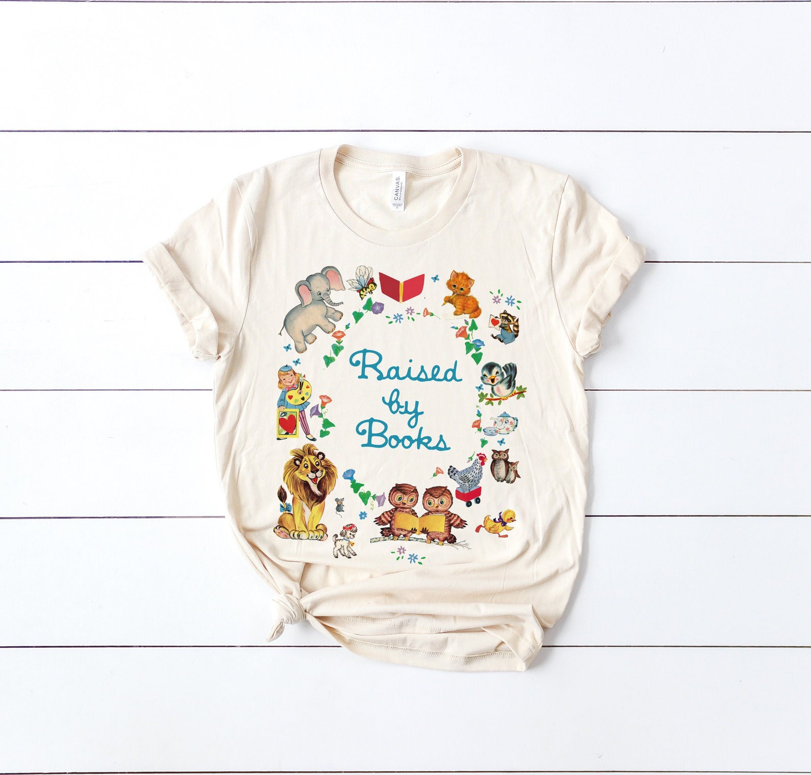 Image of a white shirt on a white paneled background. The shirt reads "Raised by Books" and features images from Little Golden Books. 