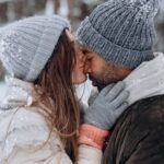 a photo of a couple outside by a forest in the snow. A white woman kisses a Black man on the nose.