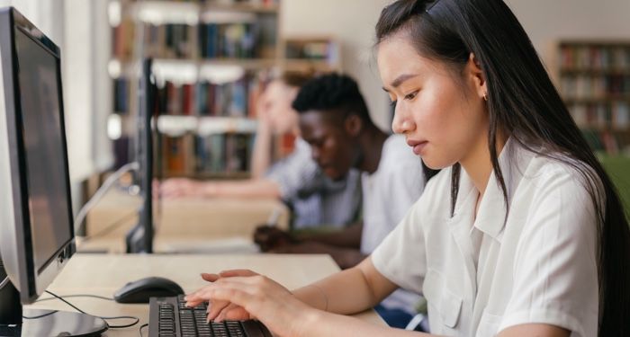 a person working at a desktop computer in a library, two more people doing the same in the background