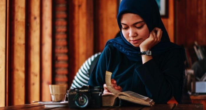 light tan skinned woman reading in hijab, camera and book in front of her on a table