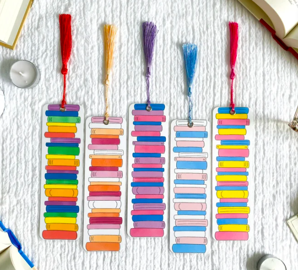 Five bookmarks with book stacks in the colors of the rainbow, trans pride flag, bisexual pride flag, and others, all with colorful tassles