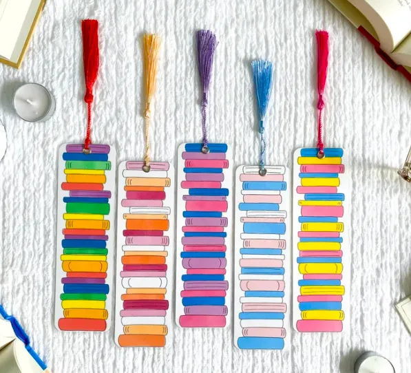 Five bookmarks with book stacks in the colors of the rainbow, trans pride flag, bisexual pride flag, and others, all with colorful tassles