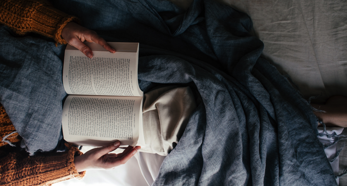 arms and legs of an unseen person reading a book in bed with a blue blanket