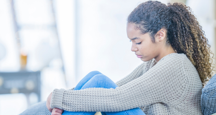 a photo of a Black teenager with long hair holding their knees and looking pensive