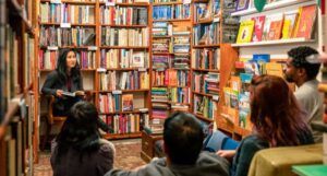 people of different skin tones are gathered around an Asian woman with a book in her hand. They are in a small, book, filled room