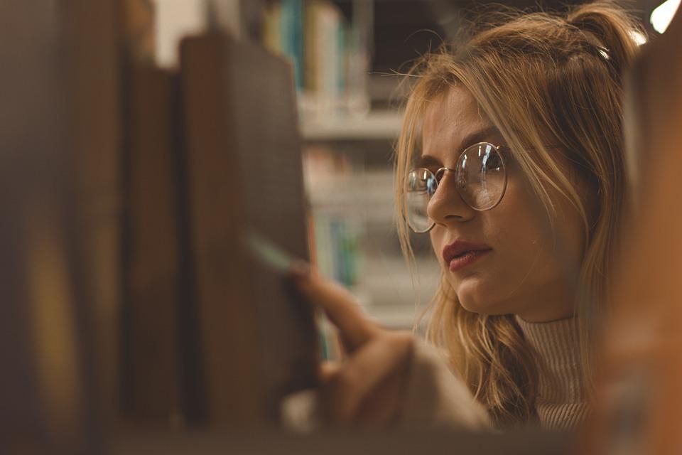 A white person with long blonde hair and glasses is browsing along a library shelf